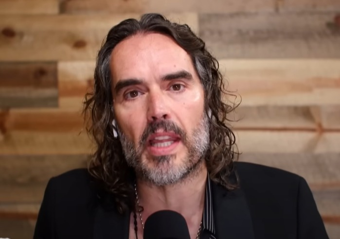 Russell Brand speaking in front of a wooden wall.