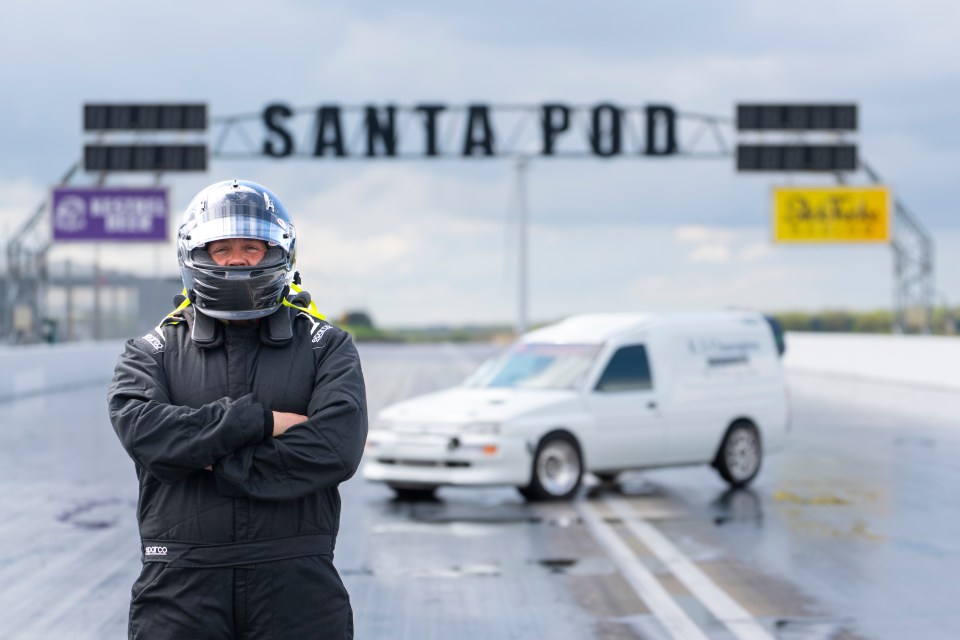 A driver in a racing suit and helmet stands in front of a modified Ford van at Santa Pod Raceway.