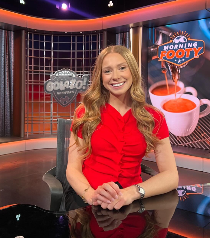 Woman in red shirt sitting at a desk in a TV studio.