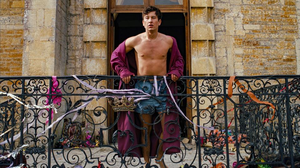 A man in a robe and shorts stands on a balcony, looking out.