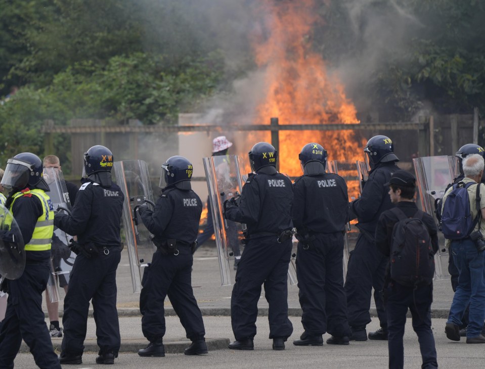 Police officers in riot gear face a burning structure during a protest.