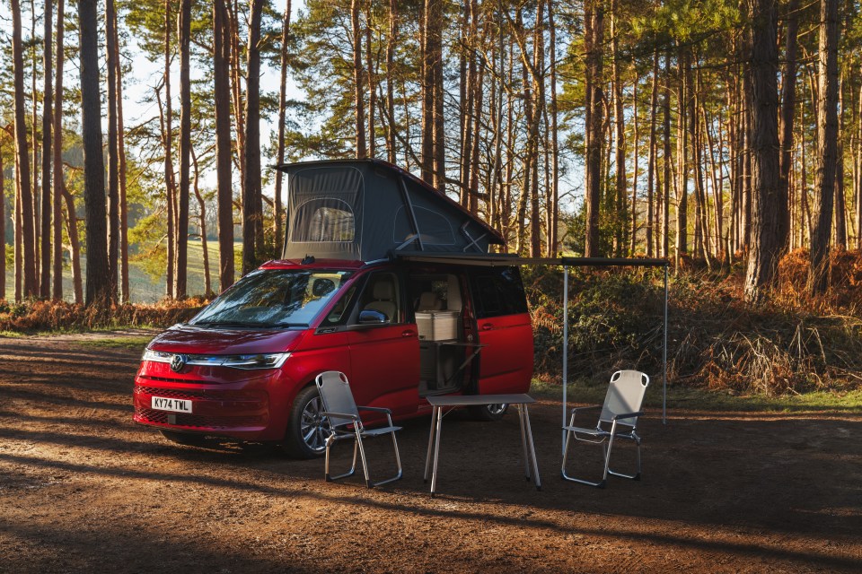 Red plug-in hybrid campervan parked in a forest with camping chairs and table set up.