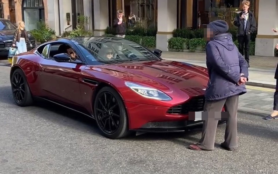 An older woman stops a red Aston Martin DB11 supercar in the street.