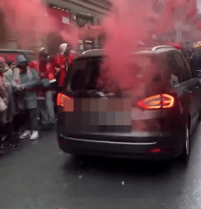 A dark-colored minivan driving through a crowd of Liverpool fans amidst red smoke.