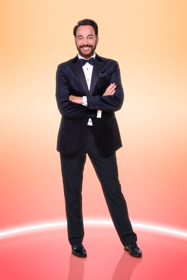 Craig Revel Horwood in a tuxedo, arms crossed, smiling.