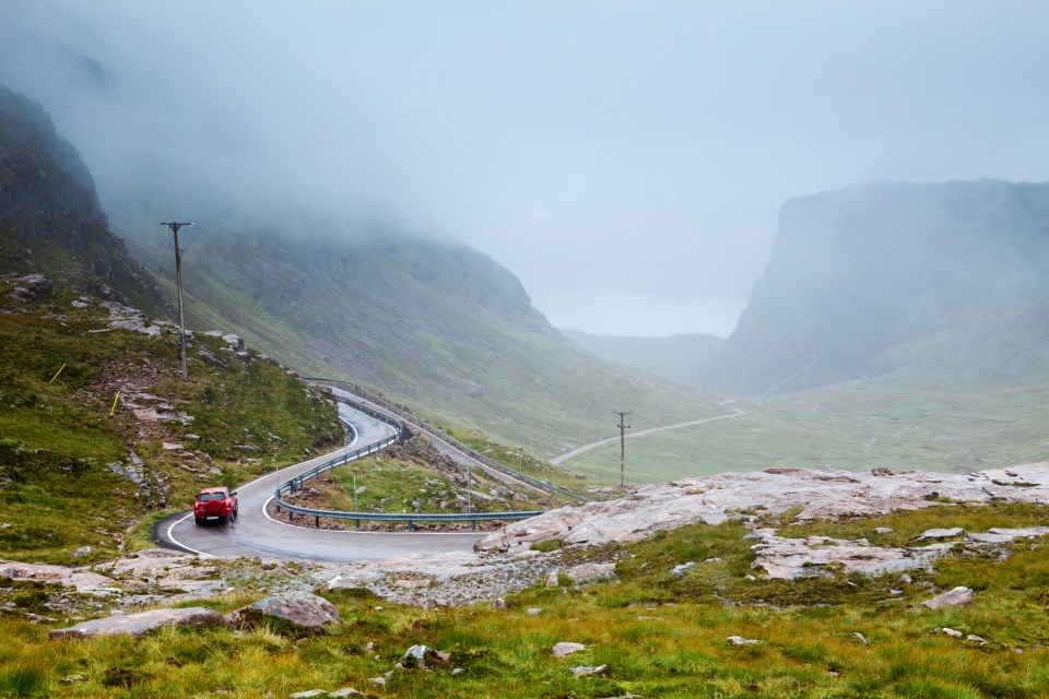 Red car driving on a winding mountain road in foggy conditions.