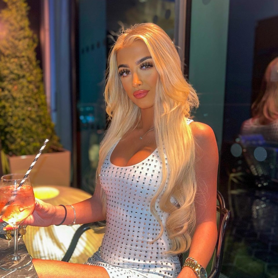 Woman with long blonde hair sitting at a table, holding a drink.