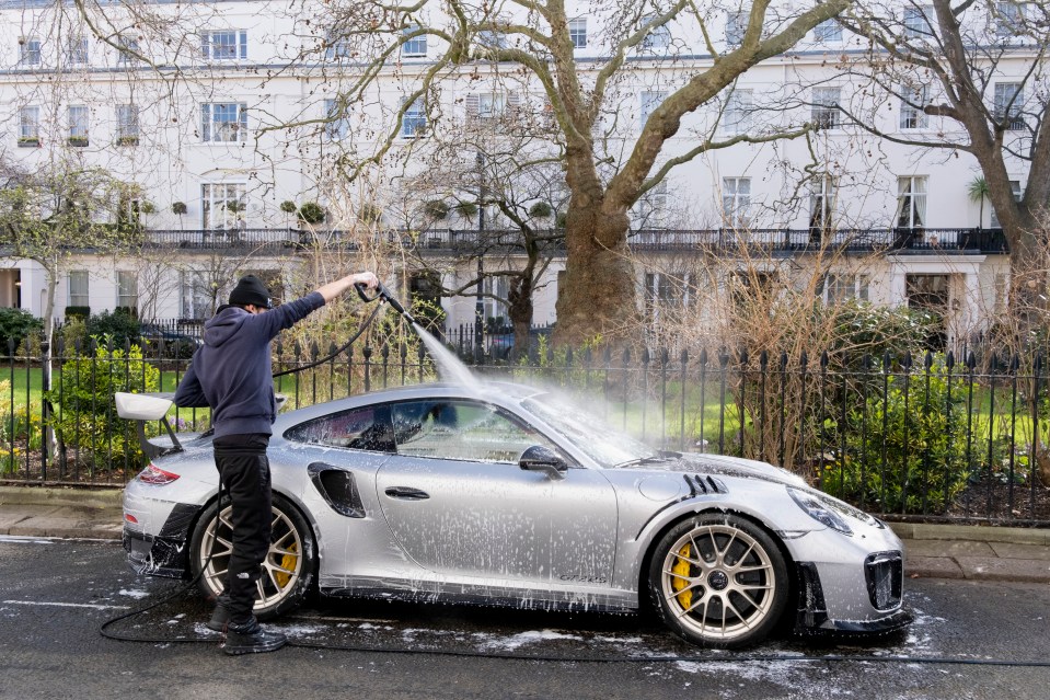 A man pressure washes a silver Porsche.