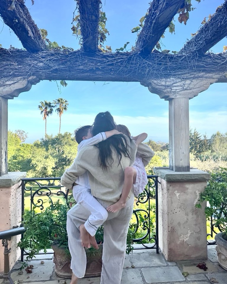 A woman holding her two young children on a balcony overlooking a garden.