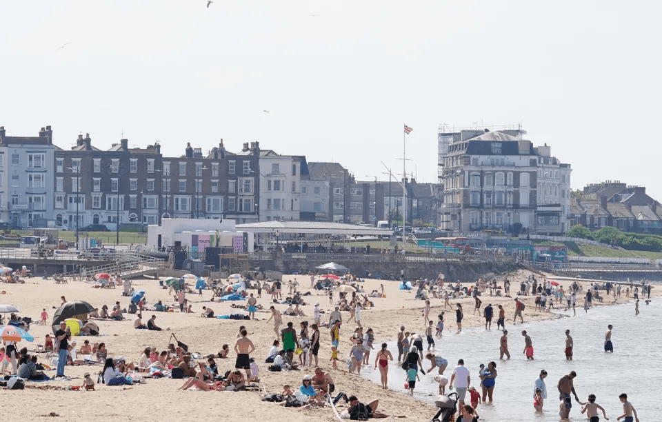 Crowded beach with people swimming and sunbathing in front of buildings.