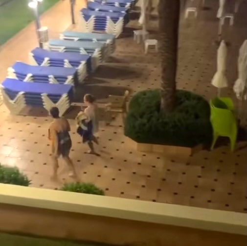 Video still of two women taking towels from sunbeds at a hotel pool at night.
